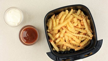 Grande plano visto de cima do cesto de uma airfryer cheio de batatas fritas douradas; duas taças com molhos ao lado